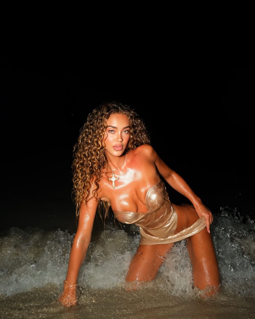 Женщина с длинными вьющимися волосами стоит на коленях в морской пене ночью, одетая в короткое золотое платье без бретелей и с крестиком на шее.